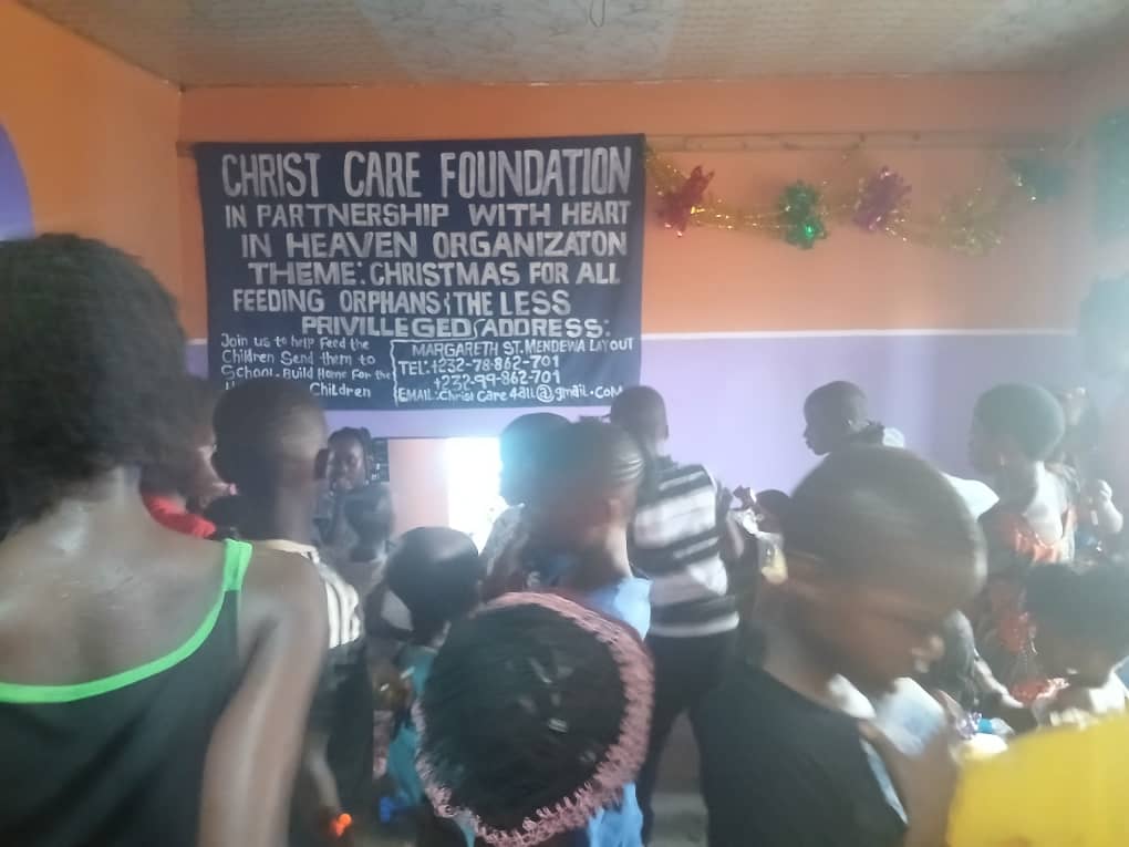 Children and volunteers gather at a Christ Care Foundation Christmas for All event in Bo City, Sierra Leone, beneath a banner recognizing the partnership with Heart In Heaven