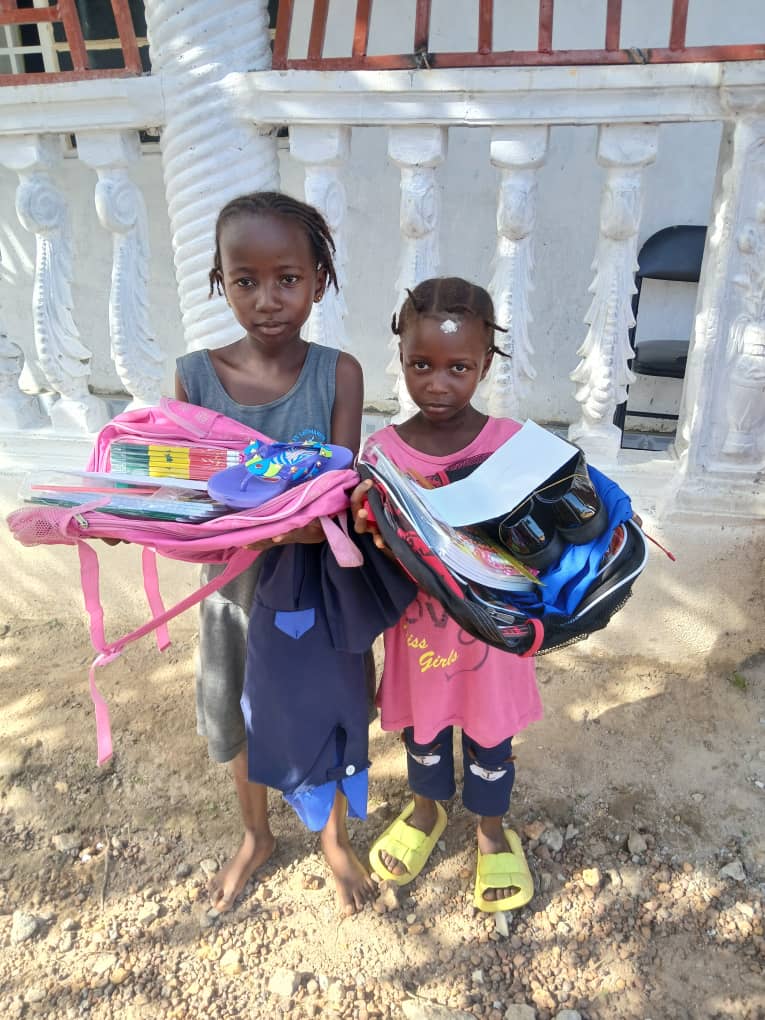 Two girls in Bo City holding school uniforms, backpacks, books, footwear, and school supplies funded through Heart In Heaven support